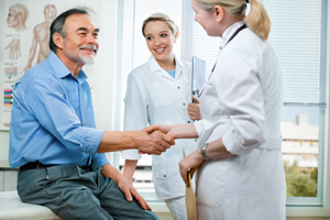 Two female Medical professionals are in a patient exam room with a male patient, smiling